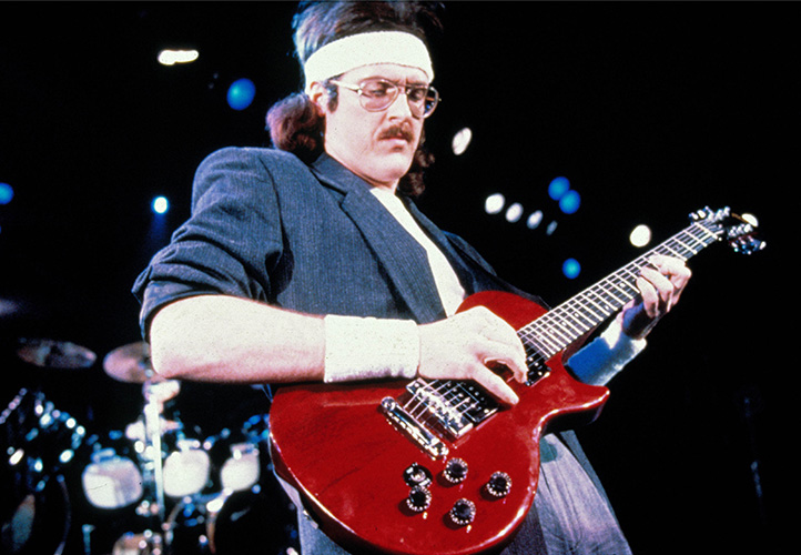 Photo of a man with a white headband on, playing a red guitar.