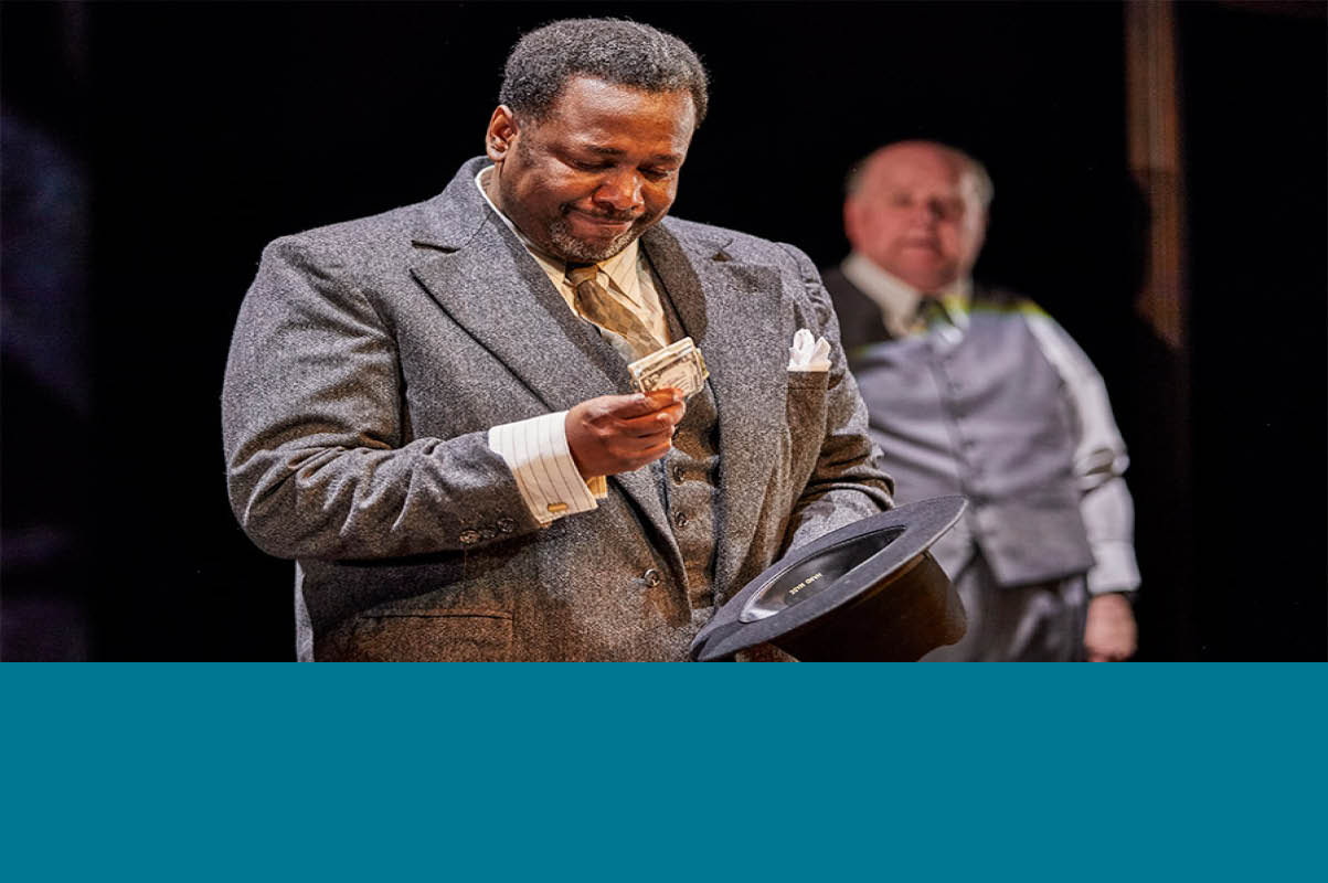 Theatrical scene of the play Death of Salesman featuring an actor wearing a grey suit looking at a dollar not with a smile. Another actor in the background also in a suite looking at him. Photo credit © Brinkoff Moegenburg