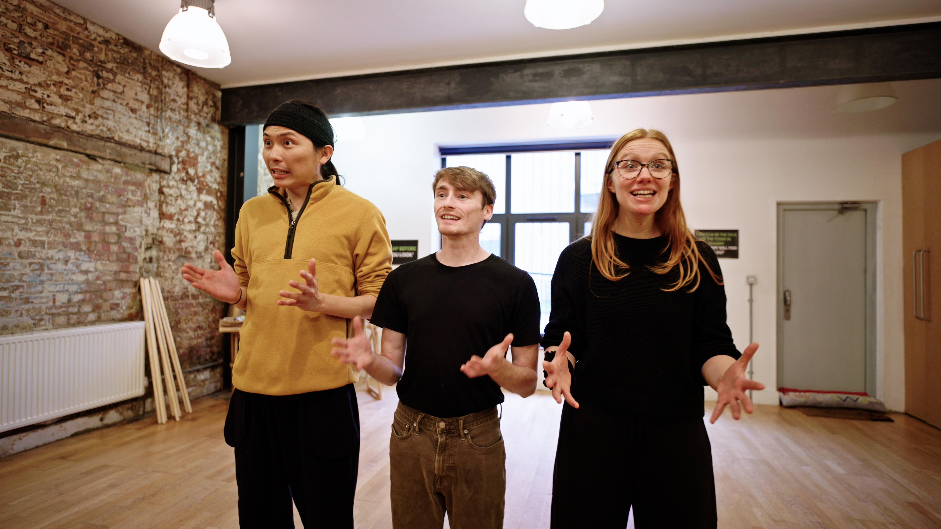 Devised Theatre Workshop: Told by an Idiot still. Two men and one woman actor stand in a row emoting with their hands spread wide.