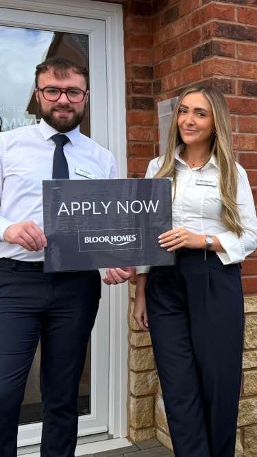 The image shows a man and a woman standing in front of a brick building, with the woman holding a sign that says "APPLY NOW".