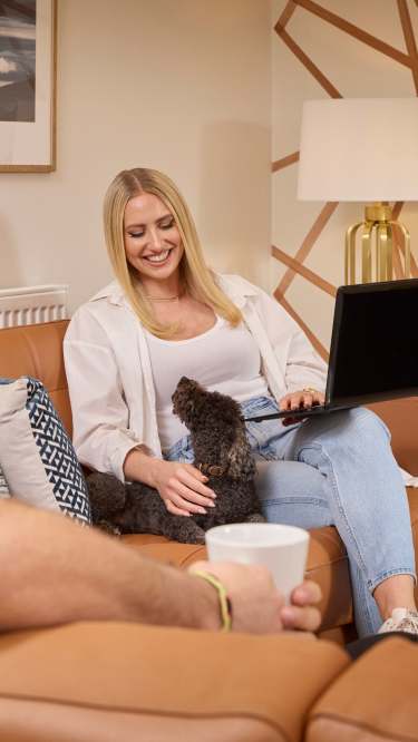 A man holding a mug sits on a leather couch in the foreground, while a woman smiles and pets a small dog on her lap as she uses a laptop in the background, with framed art and a lamp visible on the wall.