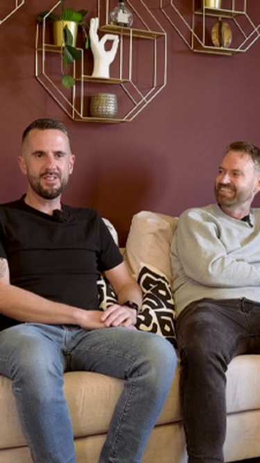 Two men sit on a cream-colored sofa with decorative pillows in front of a maroon wall adorned with geometric shelves.