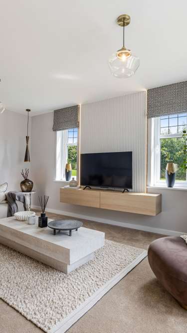 A modern living room features a plush sectional sofa and a large, light-colored coffee table in the foreground, while a wall-mounted television and two windows with patterned blinds adorn the background.
