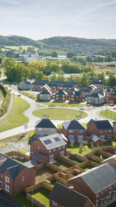 Newly built brick houses with fenced yards fill the foreground, while a circular road leads to more homes and a green, tree-covered landscape in the background.