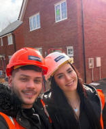 A smiling couple in hard hats and safety vests pose for a selfie in front of a new brick house under construction.