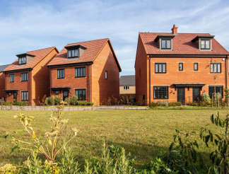 A row of modern, red-roofed townhouses surrounded by a grassy field and lush vegetation, set against a clear blue sky.