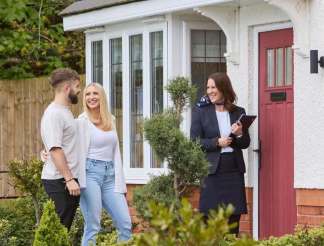 A young couple smiles at a real estate agent outside a house with a red door and bay windows, surrounded by lush greenery.