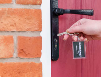 A hand holds keys to a red door, with a brick wall and doorbell in the foreground.