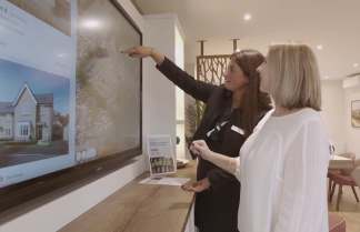 A real estate agent points to a large screen displaying property images to a potential buyer in a modern showroom.