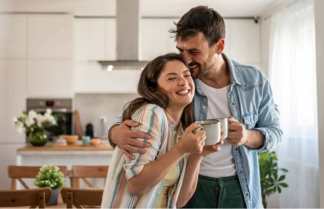 A laughing couple embraces in a bright kitchen, with a bouquet of flowers on a table in the foreground and a modern stove and cabinets in the background.