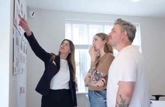 A woman in a suit points to a wall of photos while a couple looks on, with a window and blinds in the background.
