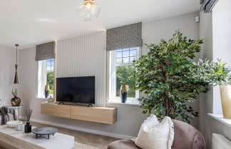 A large sectional sofa and a modern coffee table fill the foreground of a living room, while a television mounted on a wooden cabinet and windows overlooking greenery are in the background.