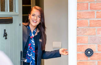 A woman in a suit and scarf smiles and opens a light blue door, with a brick wall and doorbell visible to the right.