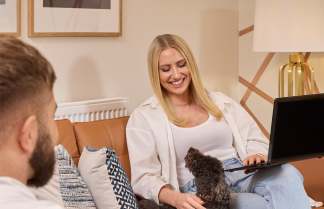 A man holding a mug sits on a leather couch in the foreground, while a woman smiles and pets a small dog on her lap as she uses a laptop in the background, with framed art and a lamp visible on the wall.