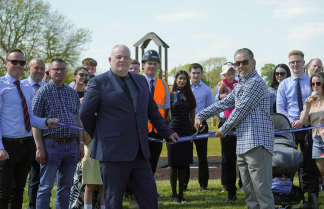 A group of people are gathered for a ribbon-cutting ceremony in front of a playground structure and new houses under a clear sky.