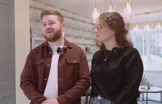 A man with a beard and a woman with glasses stand in a modern living room with a patterned wallpaper and a chandelier.
