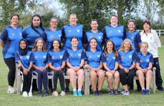 A group of women in blue cricket uniforms pose for a photo on a grassy field with a white fence and trees in the background.