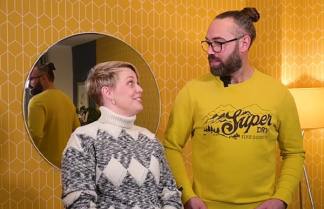 A woman in a patterned sweater and a man in a yellow "Super Dry" sweatshirt stand in front of a yellow, diamond-patterned wallpaper with a round mirror reflecting the man.