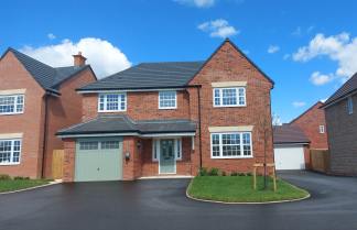 A modern brick house with a green garage door sits on a paved driveway in front of a bright blue sky with scattered clouds, flanked by similar houses.