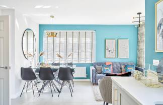 A modern dining and living area features a glass-top dining table with black chairs in the foreground, and a blue accent wall with a gray sofa and artwork in the background.