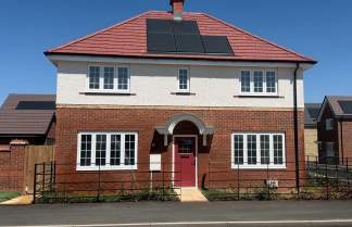 A modern two-story house with a red-tiled roof and solar panels sits behind a black metal fence, with other similar houses visible in the background under a clear blue sky.