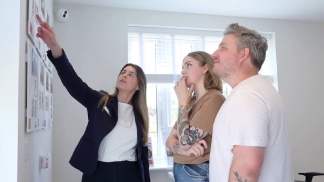 A woman in a suit points to a wall of photos while a couple looks on, with a window and blinds in the background.