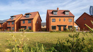 A row of modern, red-roofed townhouses surrounded by a grassy field and lush vegetation, set against a clear blue sky.