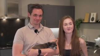 A man holds a small dog in the foreground, while a woman stands beside him in a modern kitchen with dark cabinets and stainless steel appliances in the background.