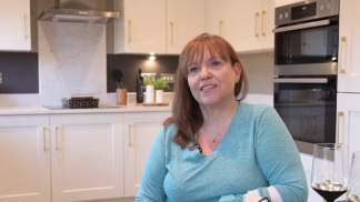 A woman smiles in the foreground of a modern kitchen with white cabinets and stainless steel appliances in the background.