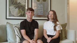 A young couple sits on a couch with decorative pillows in the foreground, while framed artwork hangs on the wall behind them.