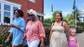 Four people, including a young child, walk along a paved path in front of a brick building and lush greenery under a bright sky.