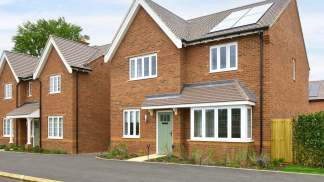 A row of modern, brick-built residential houses with gabled roofs, solar panels, and well-maintained landscaping in the foreground, set against a backdrop of a clear blue sky.
