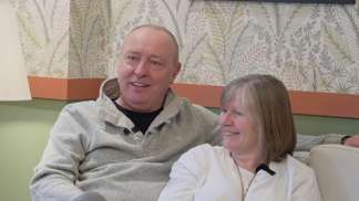 A smiling couple sits on a couch in front of a wall with a botanical pattern.