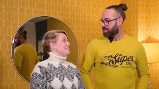 A woman in a patterned sweater and a man in a yellow "Super Dry" sweatshirt stand in front of a yellow, diamond-patterned wallpaper with a round mirror reflecting the man.