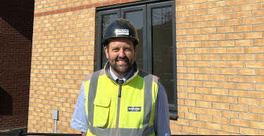 A man in a hard hat and high-visibility vest stands in front of a new, light-brown brick house with dark-framed windows, with a clear blue sky above.
