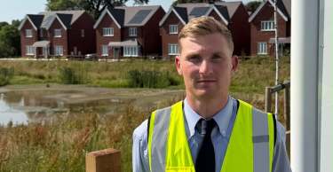 A man in a high-visibility vest stands in front of a wooden fence, with a pond and houses in the background.