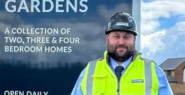 A man in a hard hat and high-visibility vest stands in front of a large sign for Bentley Gardens, with a grassy area and houses visible in the background.
