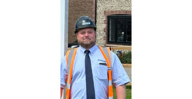 A man in a hard hat and safety vest stands in front of a brick building.