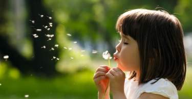 A young girl with long brown hair is blowing on a dandelion flower, surrounded by a lush green background with blurred foliage.