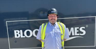 A man in a hard hat and safety vest stands in front of a dark blue construction barrier with "BLOOM HOMES" written on it, while unfinished houses are visible in the background.