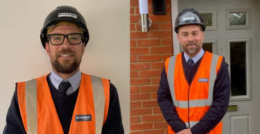 Two men in hard hats and high-visibility vests stand in front of a brick wall and a doorway.