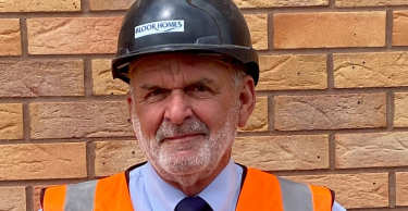 A man in a hard hat and high-visibility vest stands in front of a brick wall.