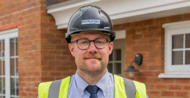 A man in a hard hat and safety vest stands in front of a brick house.