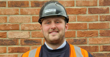 A smiling man in a hard hat and high-visibility vest stands in front of a brick wall.
