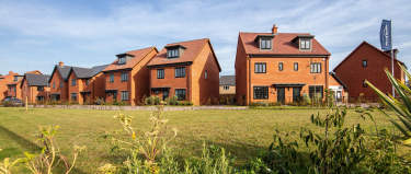 A row of modern, red-roofed townhouses surrounded by a grassy field and lush vegetation, set against a clear blue sky.