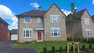 A modern stone house with a red door sits on a manicured lawn in the foreground, while other similar houses and a garage are visible in the background under a bright blue sky.