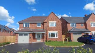 A modern brick house with a grey garage door and a blue car parked in the driveway sits under a bright blue sky with fluffy white clouds.