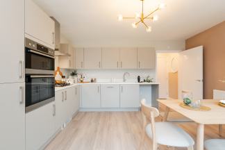 A modern kitchen with light gray cabinets and stainless steel appliances in the foreground is complemented by a dining area and a doorway to a laundry room in the background.