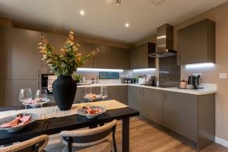 A dining table set for a meal sits in the foreground, with a modern kitchen featuring sleek cabinetry and appliances in the background.