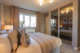 A plush bed with decorative pillows fills the foreground, while mirrored closet doors reflect the room and a window with beige curtains in the background.